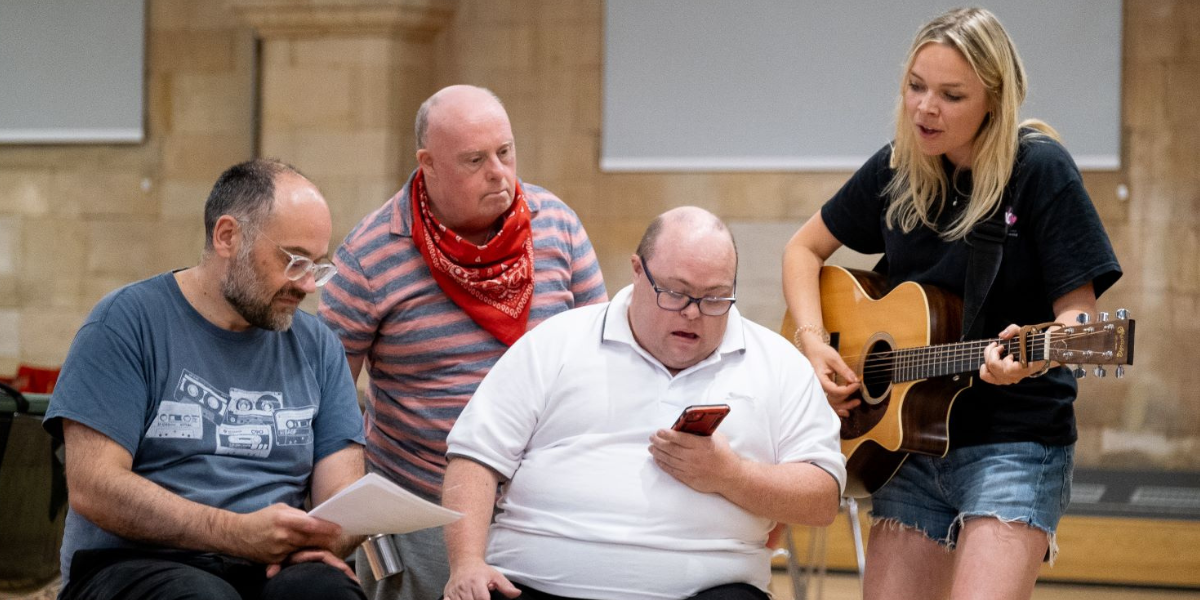 Three men sitting down and singing whilst reading a music sheet and mobile phone. A singing woman playing a guitar stands beside them.