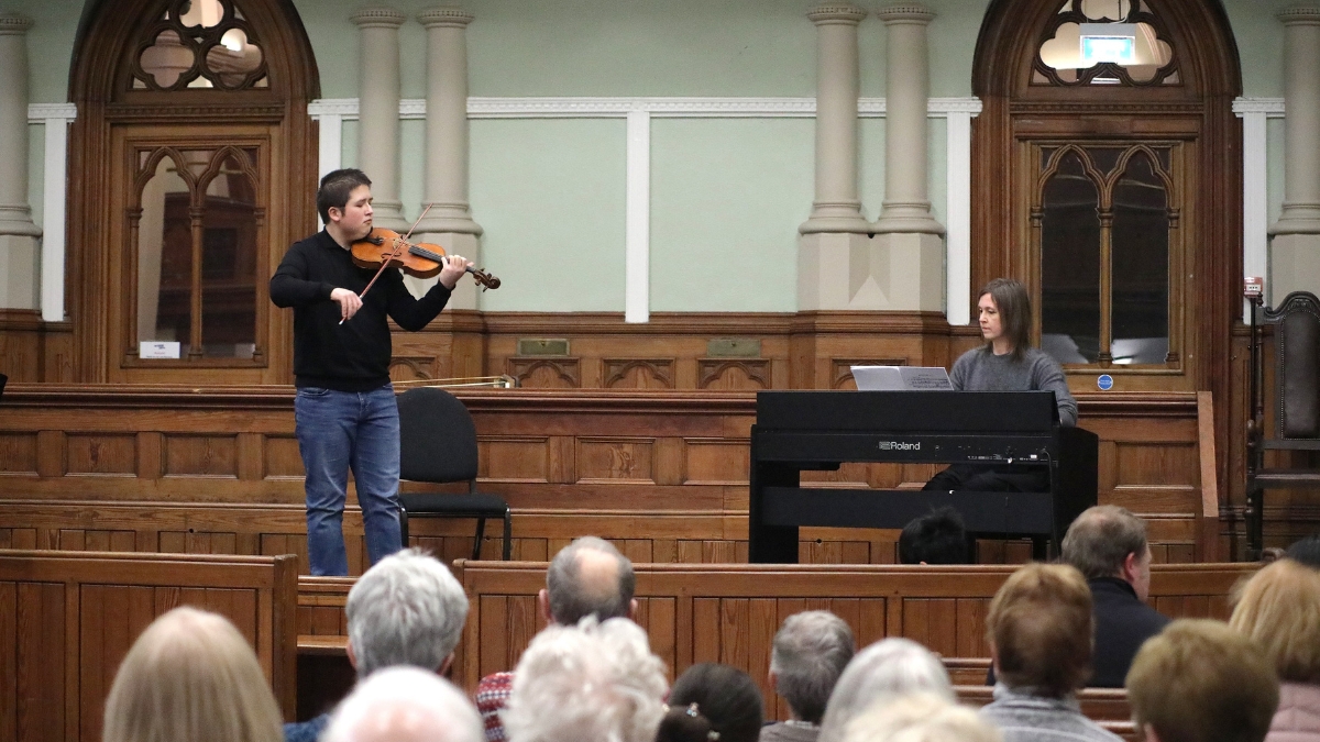A young person playing violin, accompanied by a pianist in a courtroom space, in front of an audience