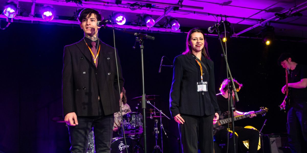 Two young people standing on a stage, with a band in the background